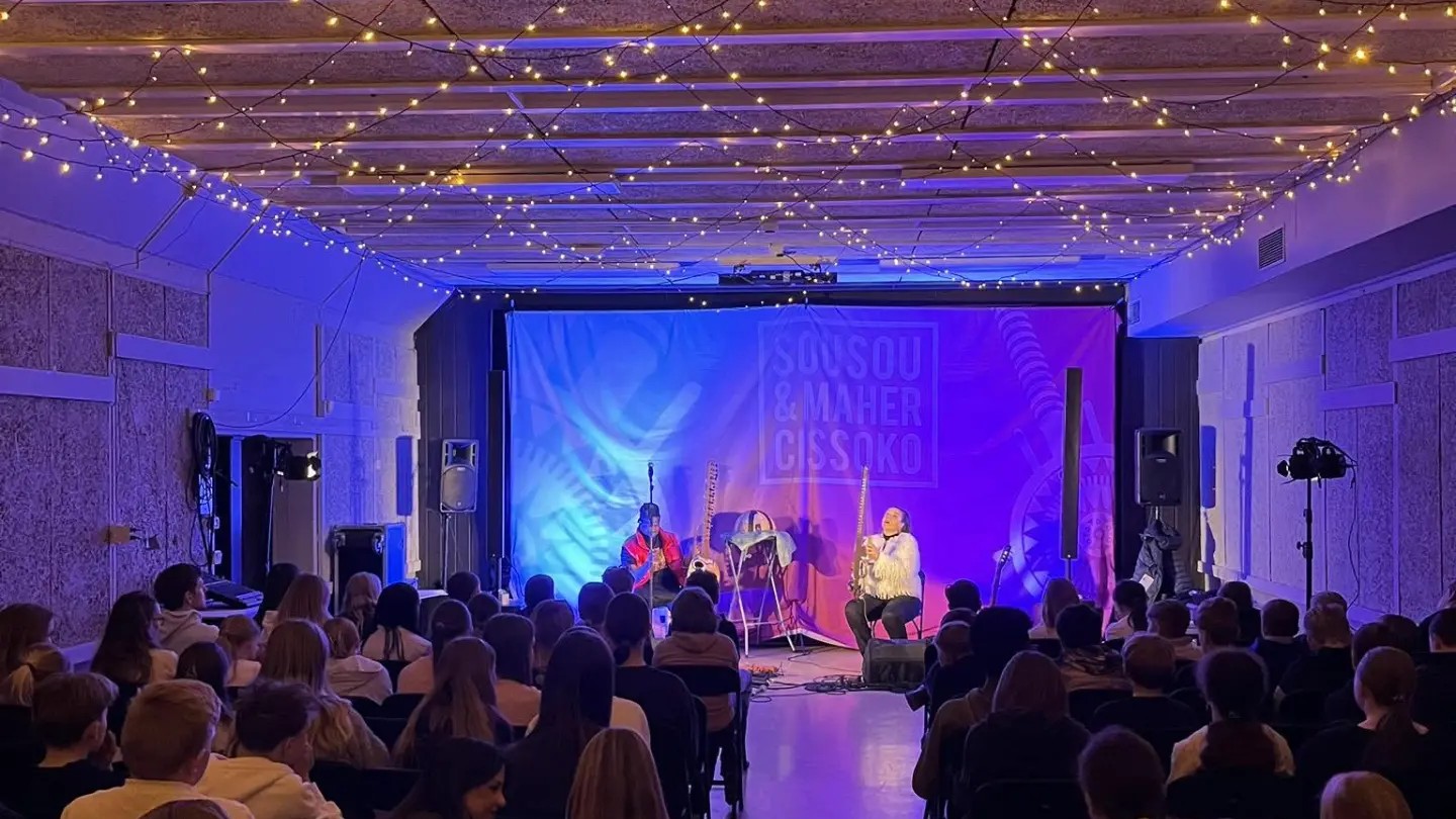 Konsert i salen på Vestfossen barneskole: elever sitter og ser to musikere spille på scenen mens lyslenker lyser i taket